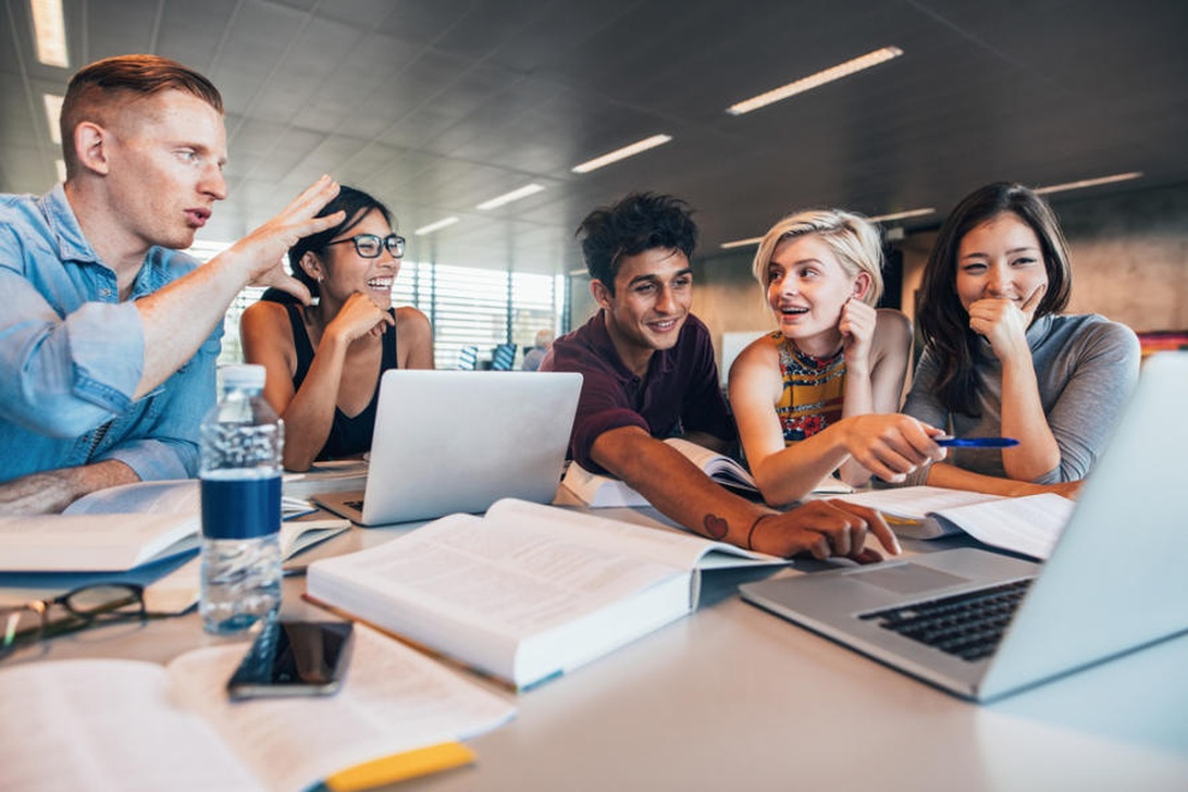 A diverse group of five young adults are gathered around a table with laptops and books, engaged in animated discussion, reflecting a positive peer learning environment.